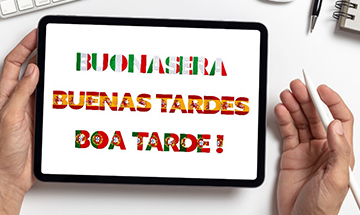 Zwei Hände halten ein Tablet, auf dessen Bildschirm die Worte „BUONASERA“, „BUENAS TARDES“ und „BOA TARDE!“